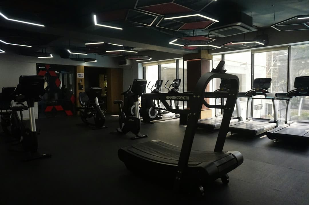 Área de cardio com esteiras e bicicletas sob teto escuro com luminárias lineares